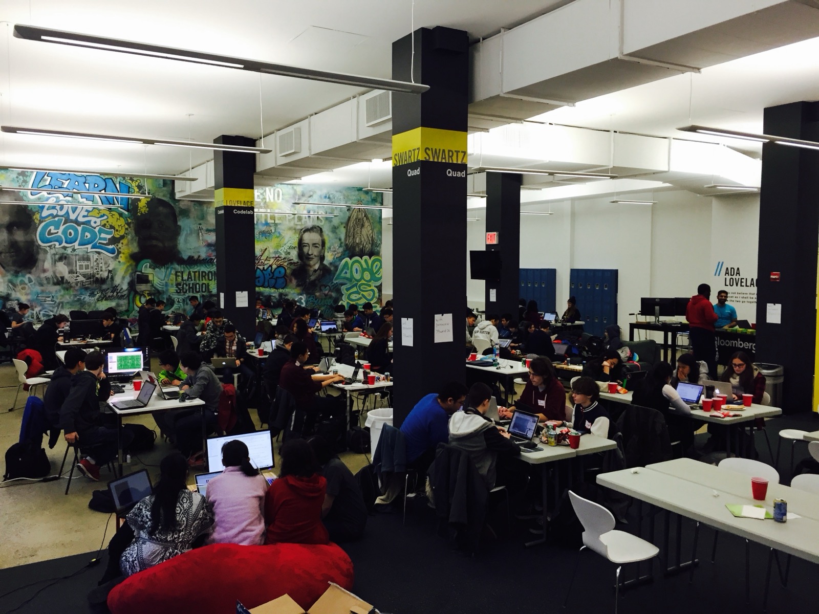 A packed classroom at Flatiron School, students at laptops beneath a mural reading Learn Love Code